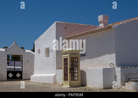 Platz mit Kopfsteinpflaster gefliest. Weiße Fassade des traditionellen Häusern. Beige Farbe der retro styled Telefonzelle. Strahlend blauen Himmel. Cacela Velha Stockfoto