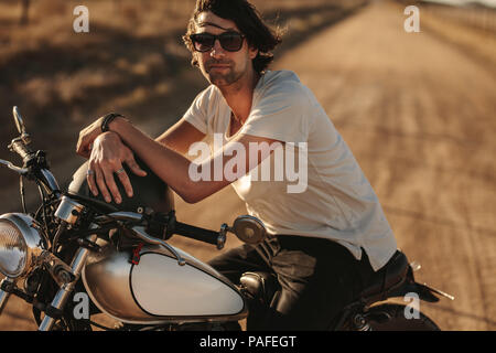 Portrait von gutaussehenden jungen Mann auf seinem Vintage Bike auf der Landstraße. Männliche biker sitzen auf seinem Motorrad und entspannt auf Landstraße. Stockfoto