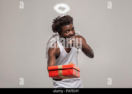 Zeigefinger Liebe und Gabe. Dunkelhäutige bärtigen African Angel lustiger Mann mit weißem Hemd, Nimbus und Winks atanding und Holding Geschenkbox. Stockfoto