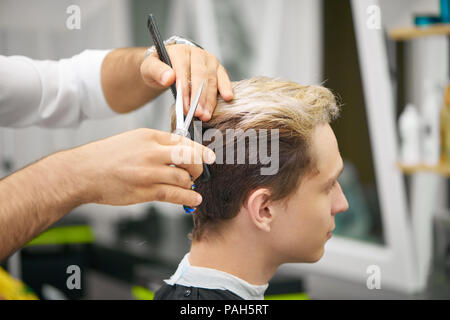 Nahaufnahme von Friseur Hände tun neuer Haarschnitt für junge Kunden. Mit schwarzem Kunststoff Kamm, metallischen scharfe Schere. Männliches Modell farbige in Haar, freuen. Arbeiten in Friseur. Stockfoto