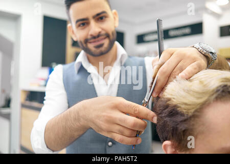 Um neue Frisur für männliches Modell mit scharfen metallische Schere. Friseur tragen klassisches weißes Shirt, grau Weste. Lächelnd, Kamera, liebevoll seinen Job. Männliches Modell in Haar getönt. Stockfoto