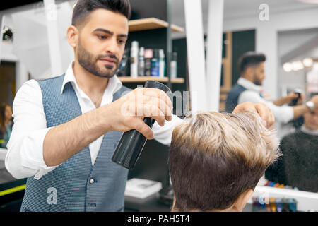 Um neue Frisur für männliches Modell mit harten Fixierung Haarspray. Friseur tragen klassisches weißes Shirt, grau Weste. Arbeiten im vorderen Spiegel im Beauty Salon. Haarpflege Produkte für den Hintergrund. Stockfoto