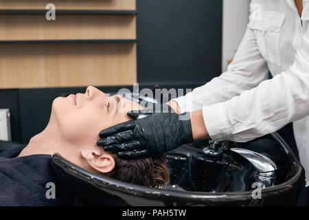 Friseur' s Hände, Haare waschen des Kunden in der beaty Salon sinken. Junge sitzt mit seinem Kopf in die schwarze Keramik Waschbecken mit geschlossenen Augen. Wohlfühlen, Relaxen. Friseur mit schwarzen Handschuhen. Stockfoto