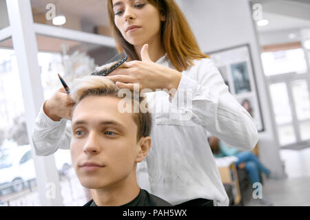 Vorderansicht des weiblichen Hairstyler tun Haarschnitt für junge Junge. Arbeiten in Spacigen beaty Salon. Männliches Modell freuen. Master mit scharfen Schere metallic und schwarz Kamm. Tragen weiße T-Shirt. Stockfoto