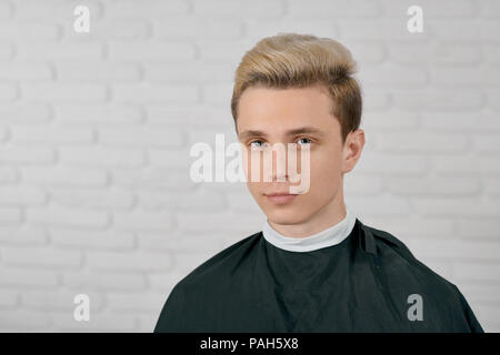 Junge männliche Modell mit blonden Haaren wartet auf neue Frisur sitzt auf weißen Hintergrund. Auf Kamera mit großen blauen Augen, dicke Augenbrauen, kleinen Ohren, nettes Gesicht. Mit schwarzem Cape abgedeckt. Stockfoto