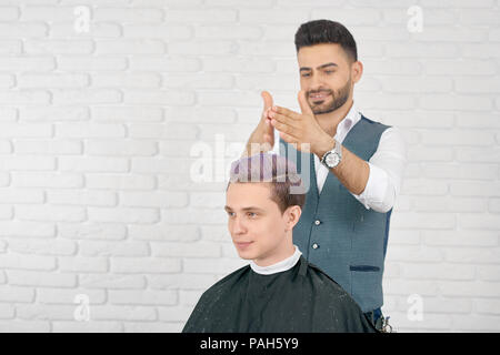 Friseur tun lila Haar Tonen für junge Kunden. Friseur legere graue Weste und Klassisches weißes T-Shirt. Kleidung Jungen bedeckt mit speziellen schwarzen Umhang. Studio sitzen auf weißem Hintergrund. Stockfoto