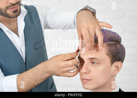 Friseur tun einen Haarschnitt für junge Kunden mit getönten lila Haare mit schwarzen Kunststoff Kamm. Tragen weiße Classic casual Shirt und graue Weste mit kleinen geometrischen Drucken. Arbeiten auf weißem Hintergrund. Stockfoto