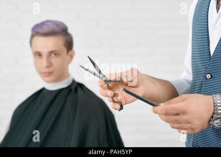 Friseur der Hände, die metallische Schere und Kamm aus schwarzem Kunststoff. Junge Kunden mit getönten lila Haar hinter und auf der Kamera sitzen. Soft Focus. Posing auf weißen Mauer Hintergrund. Stockfoto