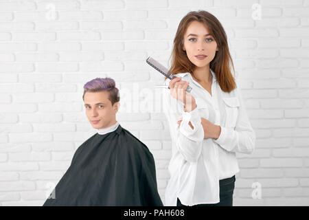 Selbstbewussten jungen weiblichen Friseur an der Kamera stand vor der Client sucht. Hairstylist, Kunststoff schwarz Kamm, scharfe metallische Schere, tragen weiße T-Shirt. Client mit getönten lila Haare. Stockfoto