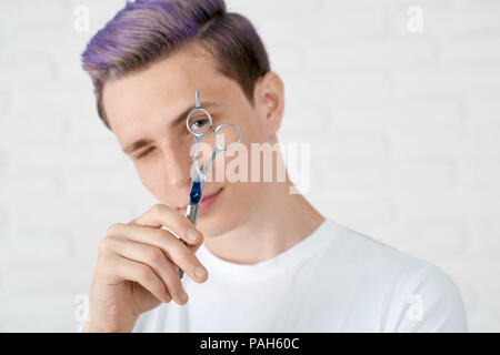 Junge hairstylist mit getönten in Violett Farbe Haare an der Kamera durch ein kleines Loch in einer scharfen Schere suchen Friseursalons. Junge tragen weiße Casual T-Shirt. Das gute Gefühl, optimistisch, suche Original. Stockfoto