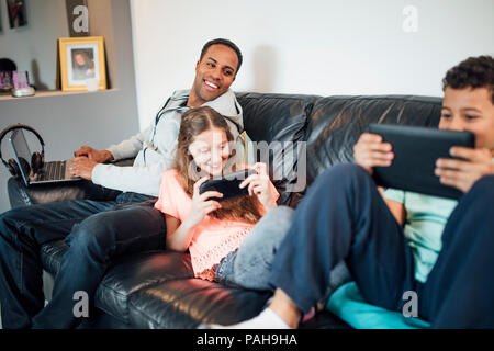 Geschwister sind, zusammen zu spielen auf dem Handheld Konsolen auf dem Sofa zu Hause, während ihr Vater ist mit einem Laptop direkt neben Ihnen. Stockfoto