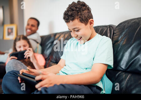 Kleiner Junge spielt auf einer Handheld-konsole zu Hause mit seiner Schwester und ihrem Vater ist sie zu beobachten. Stockfoto