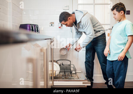 Mitte der erwachsenen Mann ist das Laden der Spülmaschine zu Hause und sein Sohn ist ihm zuzusehen. Stockfoto