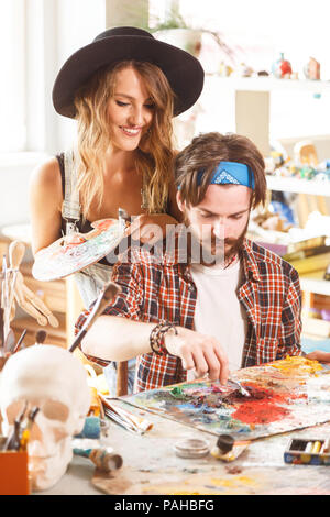 Weiblich lange Haare artist in süßen schwarzen Hut und Denim dangarees suchen Wie ihr schöner Bärtigen männlicher Kollege in Freizeitkleidung Zeichnung Stockfoto