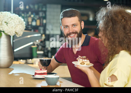 Vorderansicht des lächelnden Jungen mit einem Bart Kaffee trinken mit geschweiften Mädchen im Cafe mit modernen stilvollen Loft Interieur. Die Schale, Kuchen essen, an der Kamera schaut. Fröhlicher, positiver, Lachen. Stockfoto