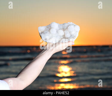 Frau Aurora Rainbow Quartz Cluster aus Indien in ihrer Hand bei Sonnenaufgang vor dem See. Stockfoto