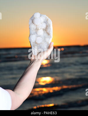 Frau Aurora Rainbow Quartz Cluster aus Indien in ihrer Hand bei Sonnenaufgang vor dem See. Stockfoto