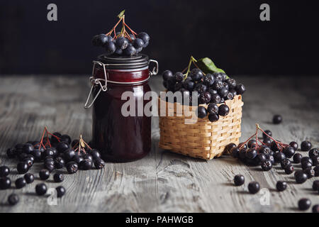 Aronia Konfitüre neben frischen Beeren. Hausgemachte aronia Marmelade im Glas Glas mit frischem Aronia Beeren auf Holztisch. Stockfoto