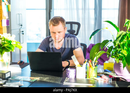 Konzentrierten jungen kaukasischen Mann in legere Kleidung Arbeiten am Laptop während an seinem Arbeitsplatz im Büro zu sitzen. Auf der Arbeit konzentriert. Freiberufler nicht Stockfoto