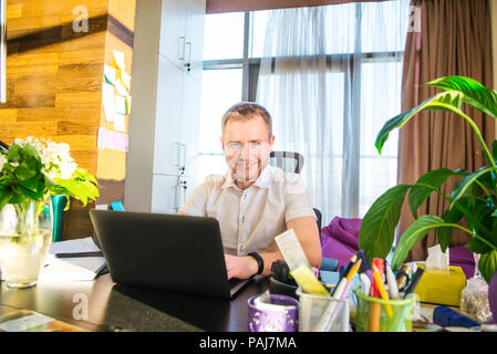 Lächelnden jungen Unternehmer von der Kamera an seinem Arbeitsplatz suchen. Erfolgreiche positiven Geschäftsmann an seiner neuen moderne Büro. Selbst Unternehmen, Freelancer wor Stockfoto