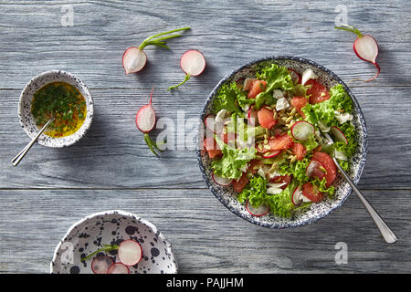 Frischen Salat mit Radieschen, Tomaten, grüne in Keramik Schüssel auf grau Holztisch. Flach. Stockfoto