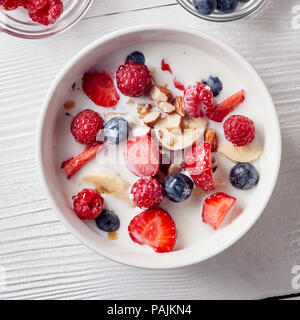 Müsli Müsli und in Scheiben geschnittenen Bananen, Erdbeeren, Beeren, gehackte Mandeln und Walnüsse mit Milch in eine weiße Keramik Schüssel auf weißer Tisch. Ansicht von oben. Stockfoto