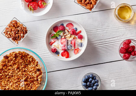 Hausgemachtes Müsli in einer Platte, Bananenscheiben, Beeren, Mandeln, Walnüsse und weiße Schüssel mit natürlichen organischen Frühstück auf weiße Holztisch. Stockfoto
