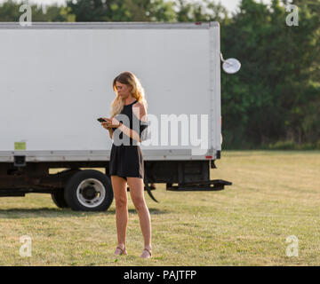 Junge Frau in einem schwarzen Jumper auf ein Mobiltelefon in einem Feld mit einer Kiste Lkw im Hintergrund Stockfoto