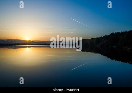 See und Stromleitungen bei Sonnenuntergang mit einer gelben Sonne am Horizont Wälder mit Bäumen auf der rechten Seite und ein Flugzeug Kondensstreifen am blauen Himmel Reflexionseigenschaften Stockfoto