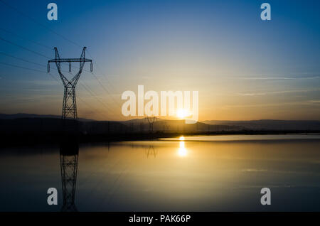 Powerlines und See bei Sonnenuntergang mit einer leuchtend gelben Sonne strahlen über die Berge und Wasser für einen Hintergrund mit einem blauen c geeignet Stockfoto