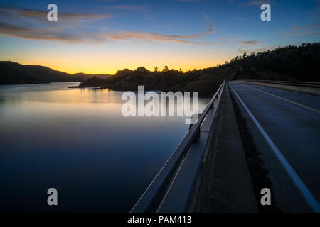 Lange Belichtung Straße und Schutzgeländer entlang einer Brücke über Don Pedro See bei Sonnenuntergang - Sierra Vorberge - Tuolumne County, Kalifornien Stockfoto