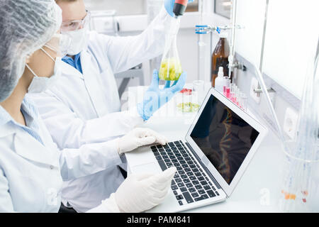 Hohen winkel Portrait von zwei moderne Wissenschaftler, die auf Essen Forschung Studium Flüssigkeiten in Becher, während am Tisch im Labor sitzen und mit Laptop Stockfoto