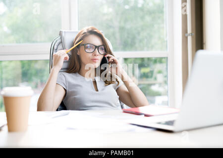 Porträt der schönen Geschäftsfrau Brille sprechen telefonisch beim Sitzen am Schreibtisch im Büro gegen Fenster Stockfoto