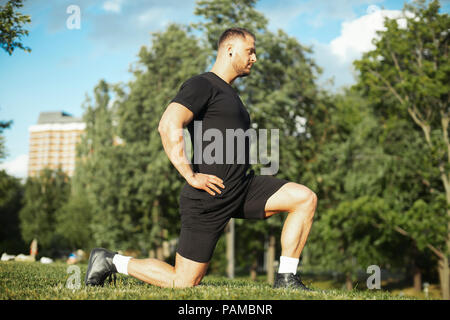 Jungen attraktiven Mann stretching Beine Ausfallschritt nach vorn im Freien tut der Mensch im Mittelpunkt und Vordergrund, Hintergrund ist unscharf. Stockfoto