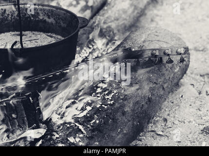 Brennende Hals der Gitarre und alten rußigen Kessel am Lagerfeuer im Wald. Schwarz und Weiß getönten Bild. Stockfoto