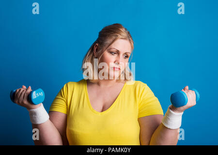 Portrait eines unglücklichen übergewichtige Frau mit Hanteln in Studio auf einem blauen Hintergrund. Stockfoto
