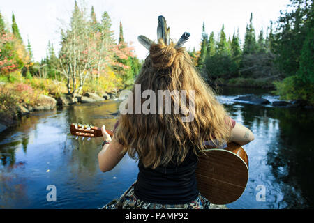 Mädchen mit Federn im Haar spielt klassische Gitarre auf einem Felsen, vor einem Fluss Stockfoto