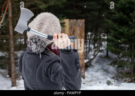 Mädchen gekleidet im Winter Kleidung hält eine Axt vor einem holzbrett als Teil einer axtwerfen Wettbewerb Stockfoto