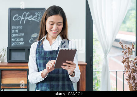 Asiatische Frau barista tragen blaue Schürze halten Tablet Computer/Menü mit lächelnden Gesicht im Cafe warten um von Kunden, Kaffee Geschäft Inhaber con Stockfoto