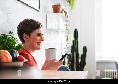 Frau sitzt in der Küche, trinken Kaffee und Kontrolle smartphone Nachrichten Stockfoto