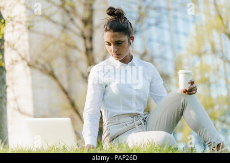 Junge Geschäftsfrau, die brechen, mit Laptop, sitzen im Gras und trinken Kaffee Stockfoto