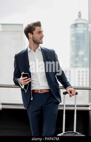 Junge Unternehmer am Fenster mit Handy, Ohrhörern und rollenden Koffer Stockfoto