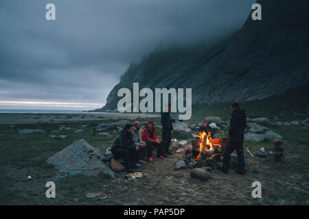 Norwegen, Lofoten, Moskenesoy, einer Gruppe von jungen Männern an einem Lagerfeuer am Strand sitzen Bunes Stockfoto