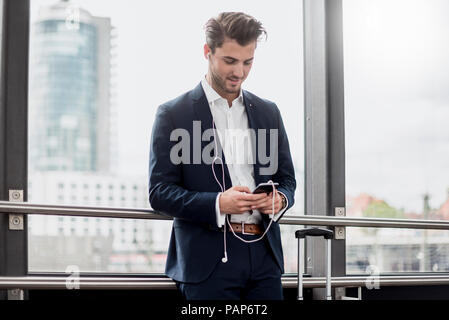 Junge Unternehmer am Fenster mit Handy, Ohrhörern und rollenden Koffer Stockfoto