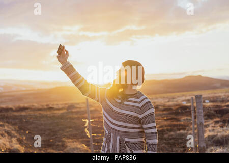 Island, junger Mann mit Smartphone, selfie bei Sonnenuntergang Stockfoto