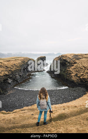 Island, Rückansicht der jungen Frau mit Rucksack und Kamera mit Blick auf Küste Stockfoto