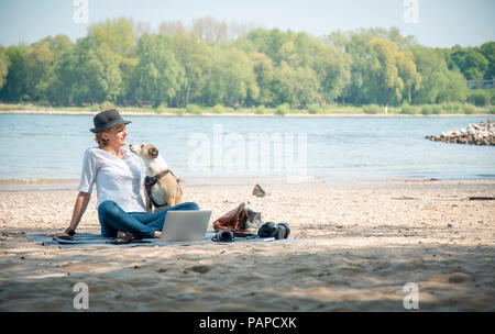 Frau sitzt auf der Decke an einem Fluss mit Hund und Notebook Stockfoto