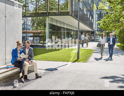 Lächelnd Kollegen mit Buch sitzt auf der Bank außerhalb der Gebäude Stockfoto