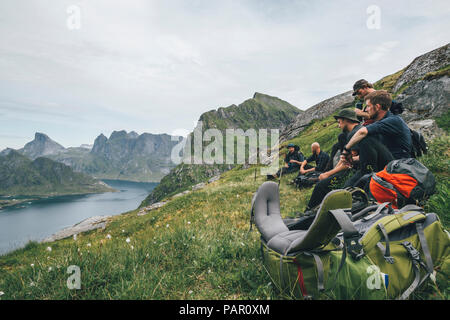 Norwegen, Lofoten, Moskenesoy, Gruppe junge Männer sitzen auf Gras, schauen über Kjerkefjord Stockfoto