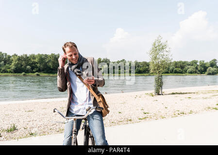 Reifer Mann mit Fahrrad mit Smartphone und Kopfhörern bei Rhein Ufer Stockfoto